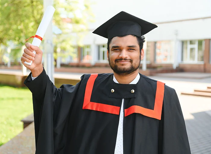A male student is standing with his graduation certificate outside the college building